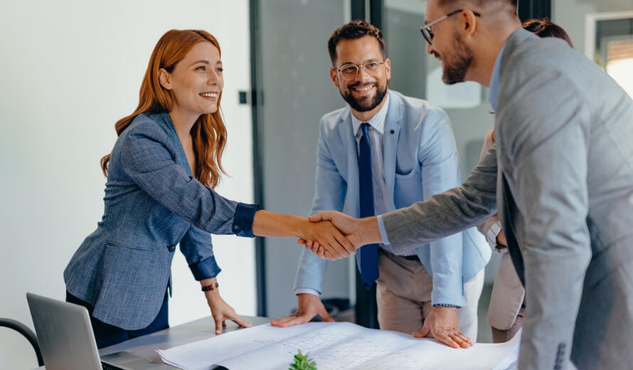 A professional business meeting with four colleagues engaged in a handshake around a table with blueprints, symbolizing successful collaboration, teamwork, and productive negotiations in a modern office setting.