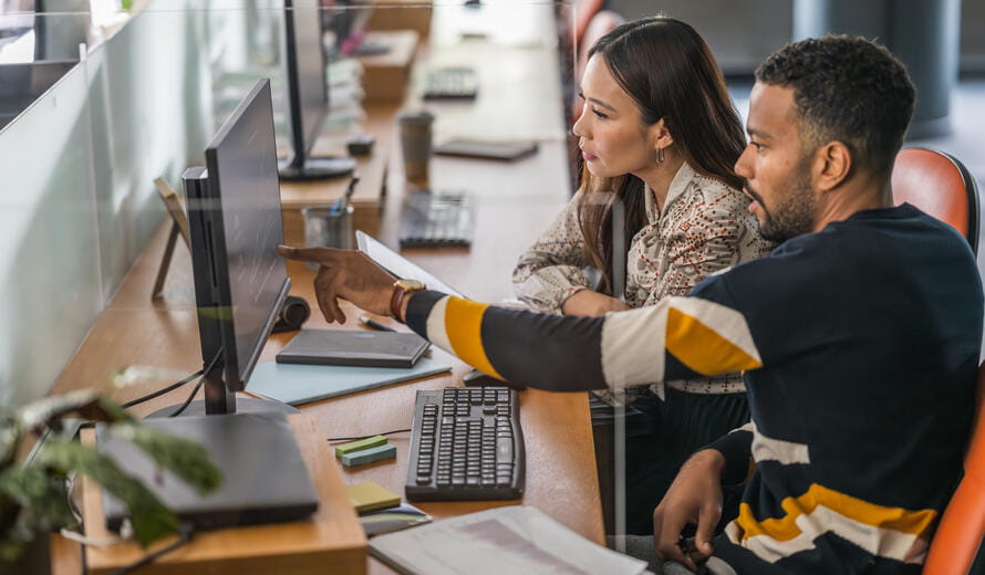 A mid adult Asian businesswoman and a mixed race businessman examine spreadsheets on a desktop PC in a well-lit, contemporary office setting, both dressed in smart casual attire.